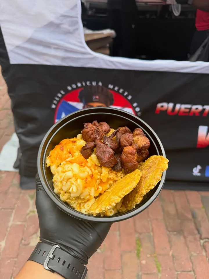 A gloved hand holding a bowl with mac and cheese, tostones and meat.