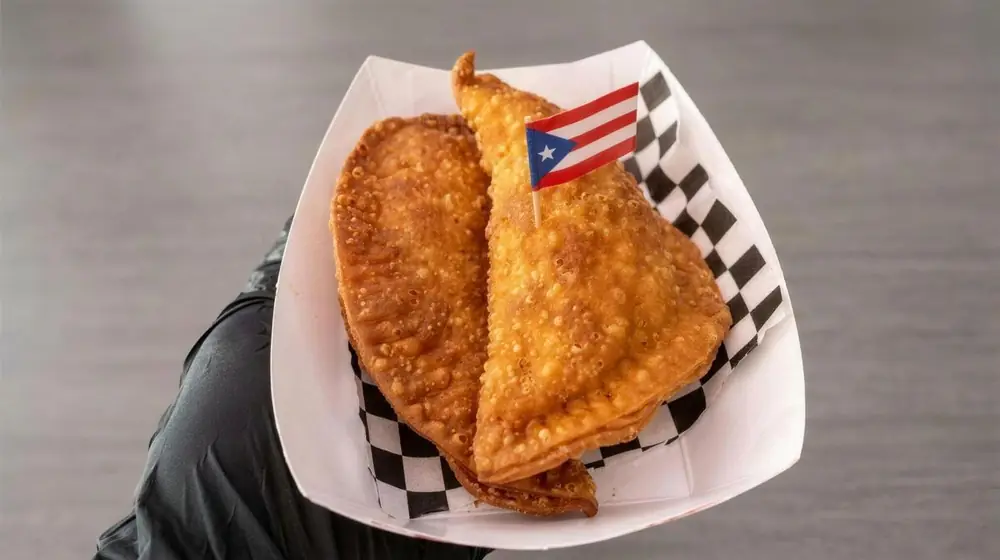 Two fried empanadas in a paper boat lined with checkered paper, a small Puerto Rican flag on a pick.