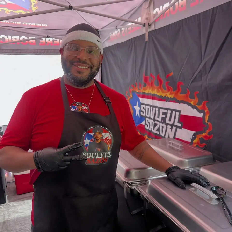 A cook in a black apron smiling and pointing at the logo, behind him the Soulful Sazón banner and steam trays.